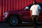 A Black man in a white t-shirt stands next to a dark red Jeep Grand Cherokee, somberly looking off to his right.