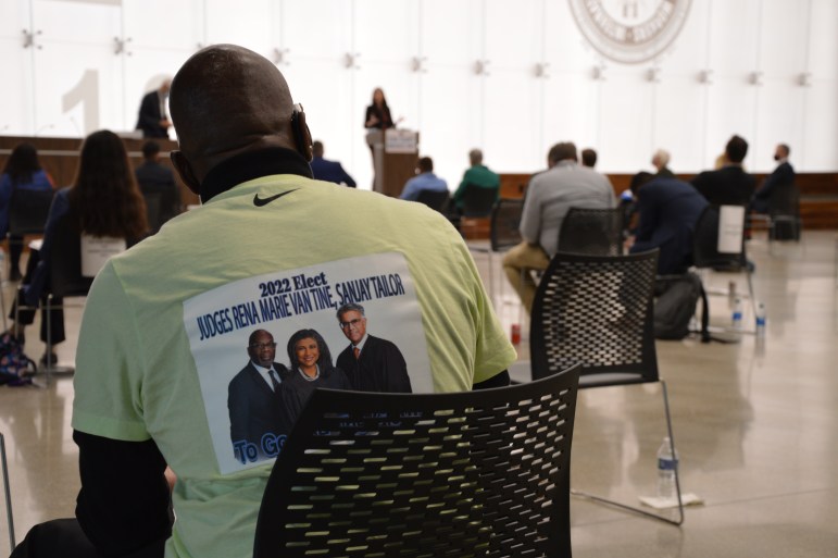 A man sits facing a podium in the background. He is wearing a green shirt that reads "2022 Elect Judges Rena Marie Van Tine, Sanjay Tailor" with a pictue of three people on the back.