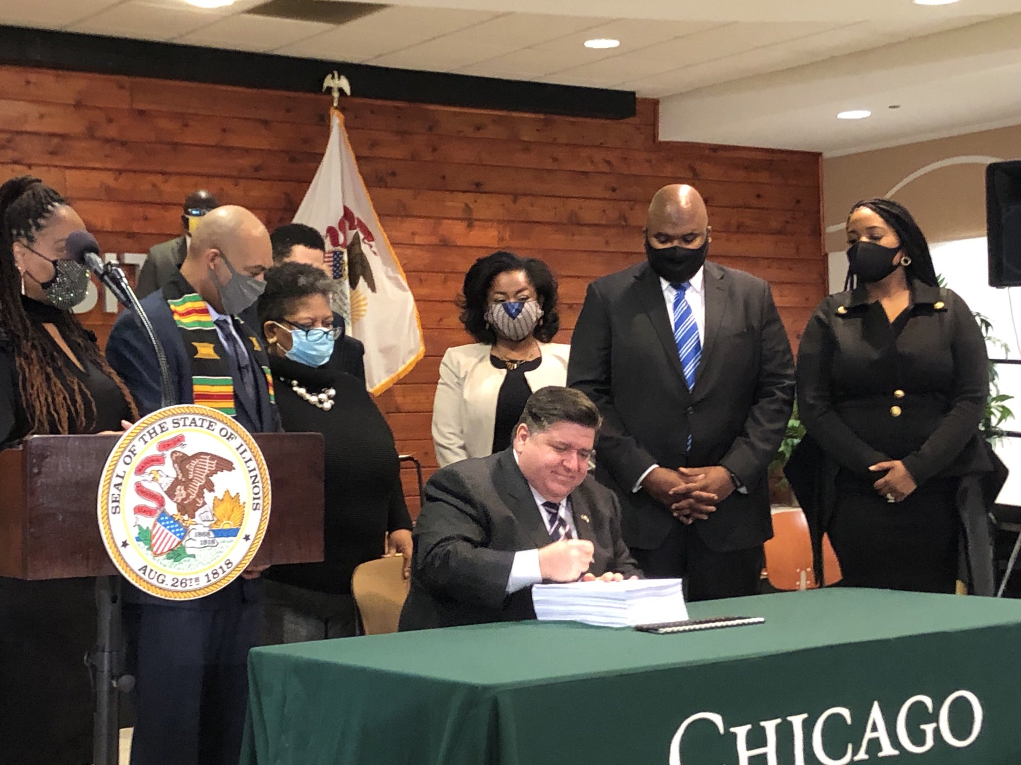 Gov. J.B. Pritzker signs criminal justice reform package sitting at a green table surrounded by lawmakers in suits and face masks..
