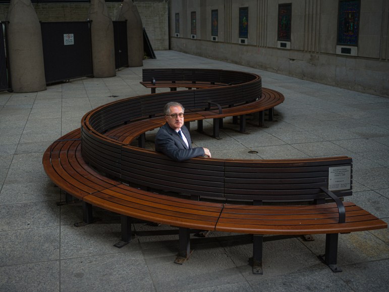 A man in a suit with glasses poses for a portrait on a large curved wooden bench in the middle of an ornate lobby.