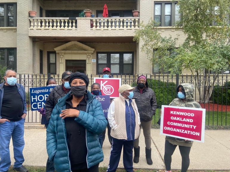A group of older Black people in coats stand outside a white brick building with signs that read Kenwood Oakland Community Organization and Lugenia Burns Hope Center. One woman stands in front with a puffy blue coat and a mask.