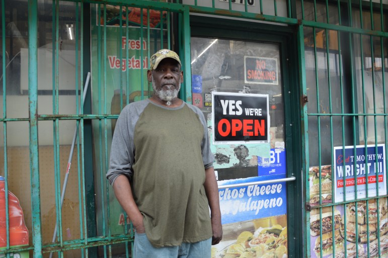 A Black man with a white beard tied in a ponytail stands in front of a store with bars on the windows and a large Yes, we're open sign.