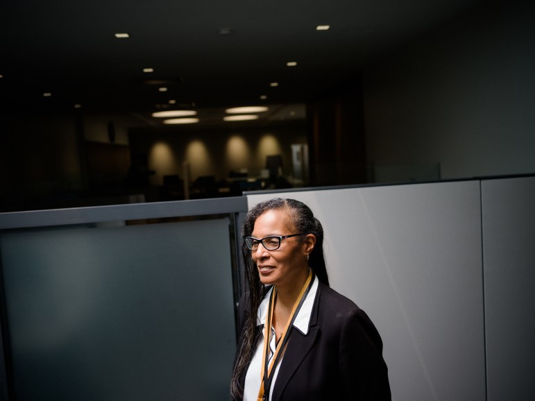 A portrait of a Black woman with long hair and glasses standing in a mostly dark and empty office.