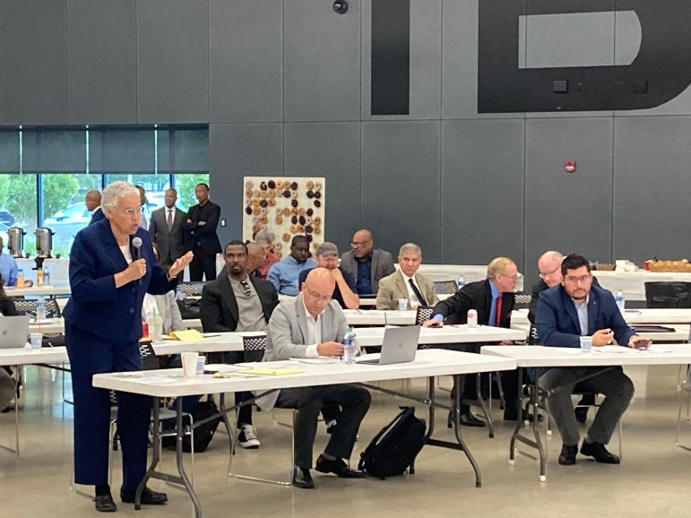 A group of mostly men in sports coats sit at rows of folding tables. On the left side, Toni Preckwinkle, a Black woman with grey hair in a navy suit, stands with a microphone in her hand to address slated candidates for judge.