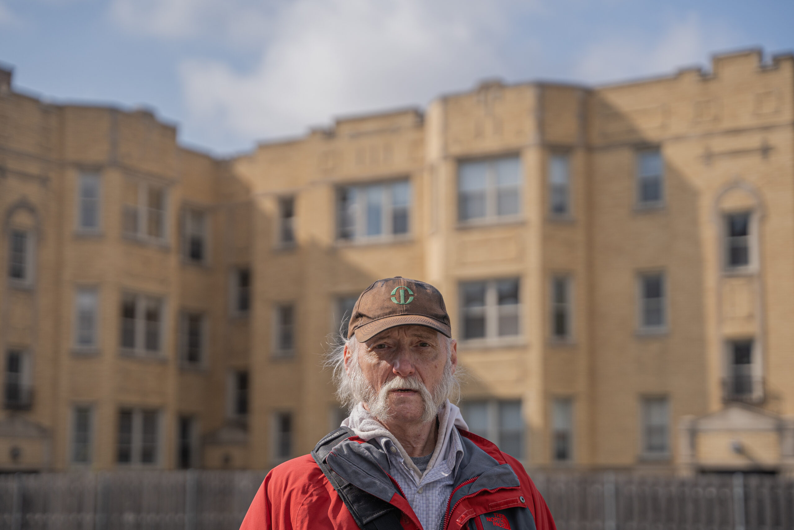 An older man with grey hair and a grey mustache stands outside in a red jacket and brown hat. There's a light brick building in the background.