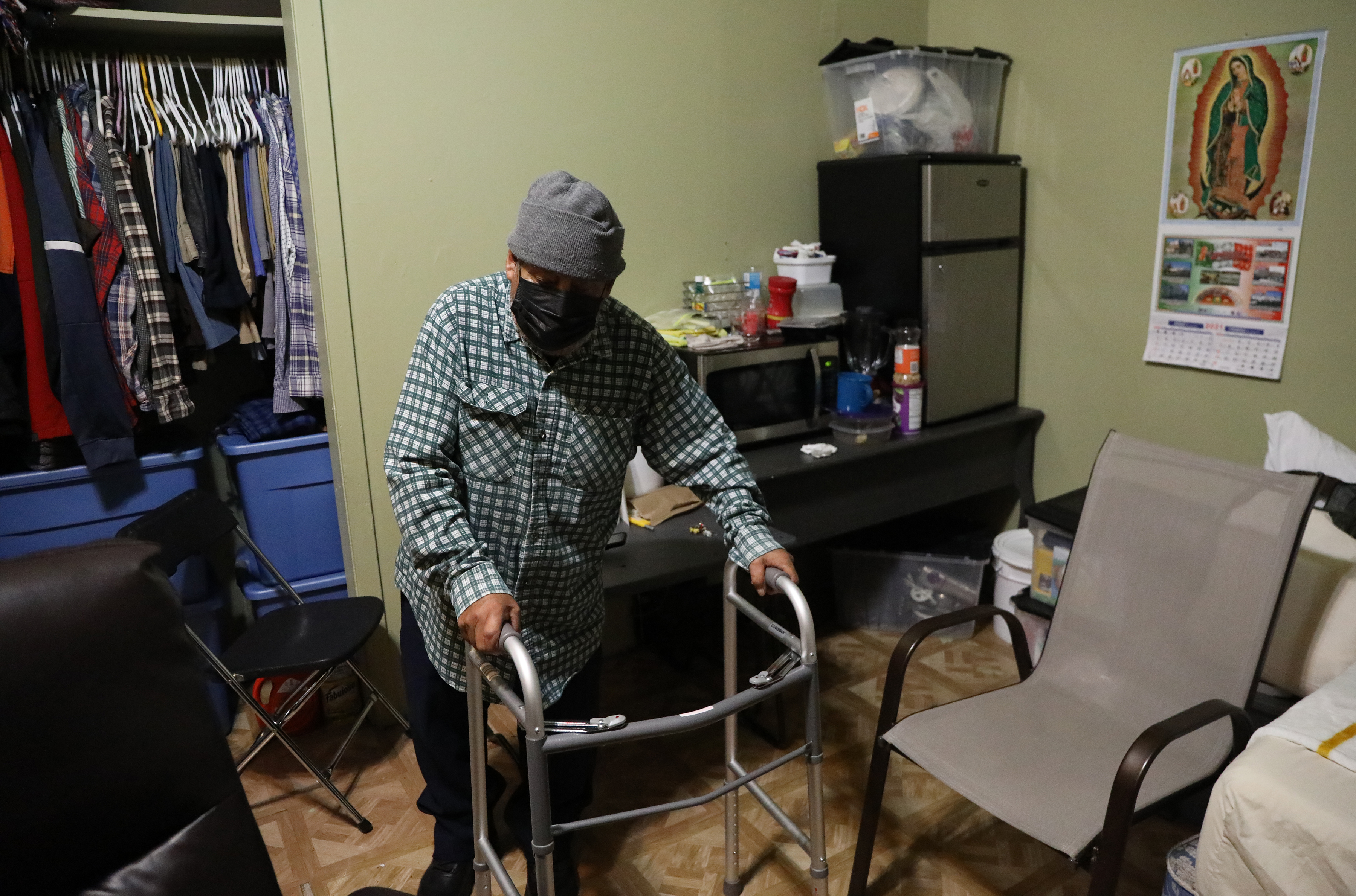 An elderly man stands with a walker in a small apartment with a few chairs, a microwave and mini-fridge, and a closet full of clothes behind him.