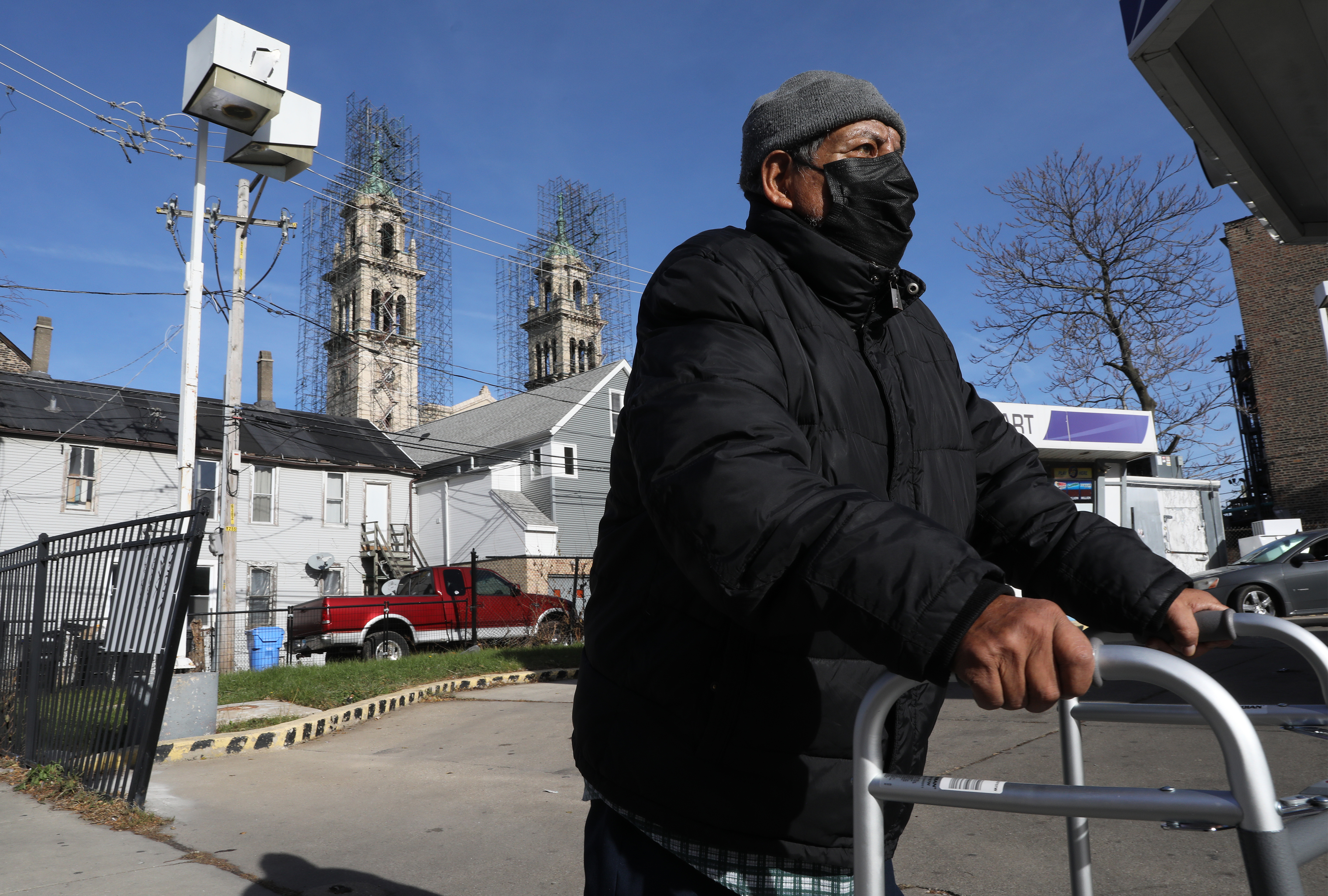 An elderly man holds a walker as he walks down the street. He's wearing a black winter coat and black face mask. Behind him are the steeples of a church against a clear blue sky.