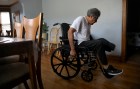 An elderly man with one amputated leg wheels himself on a wheelchair past a dining room table.