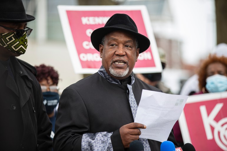An older Black man in a black hat with a salt-and-pepper goatee speaks in front of a group of people holding signs.